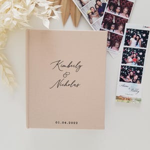 May include: A light pink linen-covered photo album with the names "Kimberly & Nicholas" and the date "01.04.2022" printed on the cover. Photo booth strips with images of people are next to the album. Decorative dried flowers and a sunburst-shaped object are in the background.