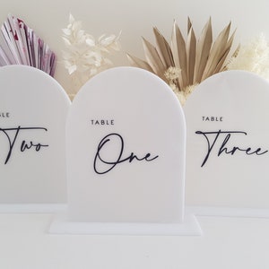 May include: Three white acrylic table number signs with rounded tops and black calligraphy writing. The numbers are "One", "Two", and "Three".