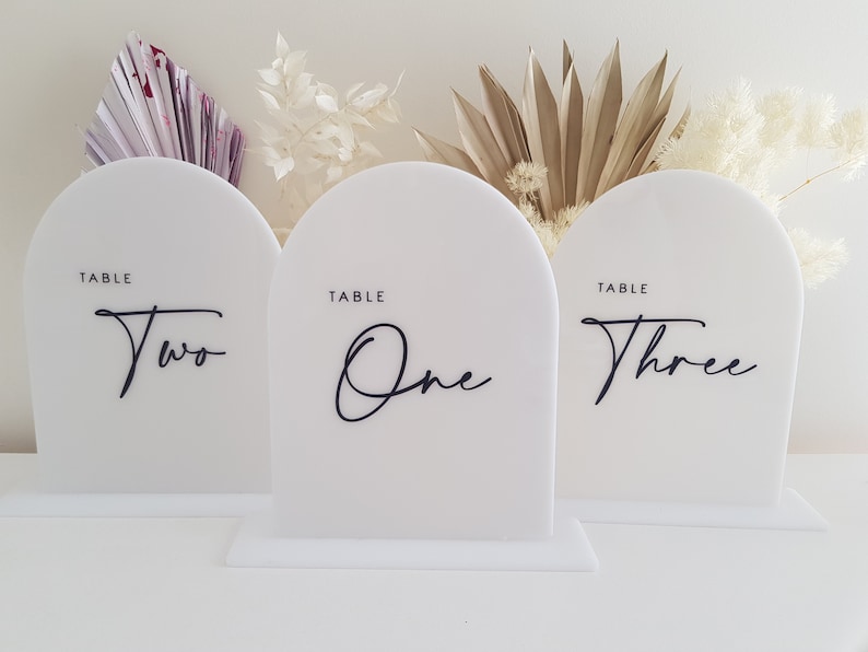 May include: Three white acrylic table number signs with rounded tops and black calligraphy writing. The numbers are "One", "Two", and "Three".