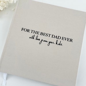 May include: A light gray journal with the text "FOR THE BEST DAD EVER with love from your kids" in black. The journal has a white ribbon bookmark and is on a white surface.