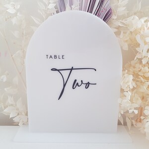 May include: White acrylic table number with a rounded top and a black base. The number "Two" is written in a cursive font with the word "Table" above it.