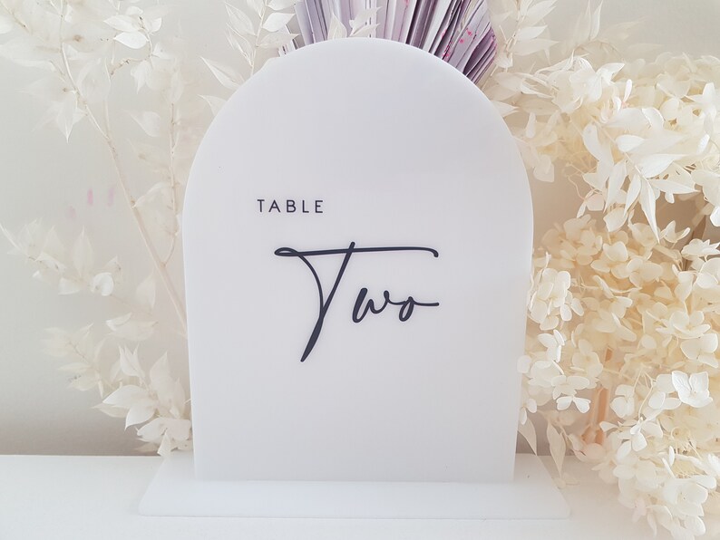 May include: White acrylic table number with a rounded top and a black base. The number "Two" is written in a cursive font with the word "Table" above it.
