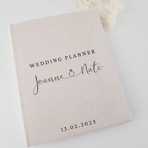 May include: A beige wedding planner with the text "WEDDING PLANNER" and the names "Joanne & Nate" in elegant script. The date "13.02.2023" is printed at the bottom. The planner has a textured cover.