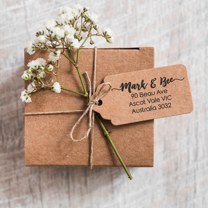 Op de afbeelding: Een bruine cadeaudoos, vastgebonden met touw en versierd met een takje witte bloemen. Een bruin cadeau-etiket is aan de doos bevestigd met de tekst "Mark & Bec 90 Beau Ave Ascot Vale VIC Australia 3032"