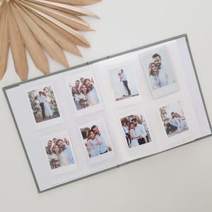 Op de afbeelding: Open fotoalbum met acht instantfoto's van stellen en groepen. Het album heeft een groene kaft en witte pagina's. Een gedroogd palmblad en witte bloemen staan op de achtergrond.