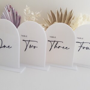 May include: Four white acrylic table number signs with rounded tops. The numbers one to four are written in black cursive script. Each sign reads "Table" above the number.