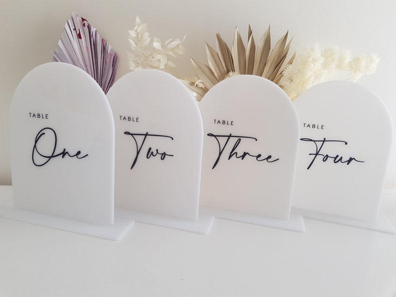 May include: Four white acrylic table number signs with rounded tops. The numbers one to four are written in black cursive script. Each sign reads "Table" above the number.