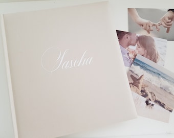 May include: A white photo album with the name "Yascha" written in cursive on the front cover. The album is surrounded by three photographs, one of a couple kissing, one of a couple holding hands, and one of a starfish on a beach.