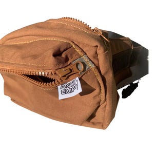May include: A tan fanny pack with a zippered main compartment and a smaller front zippered pocket. The bag has a rectangular shape with rounded edges. A small white tag with the text "ANTI EMF" is attached to the front.