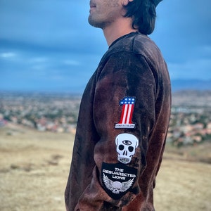 May include: A brown sweatshirt with several patches on the left sleeve. The patches include an American flag, a skull with an eye, and a skull with wings and the text "The Resurrected Lions".