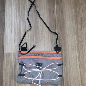 May include: A grey and orange crossbody bag with a black adjustable strap. The bag has a drawstring closure and a reflective logo with the text "24/7" and "Ultra-Trezzo".