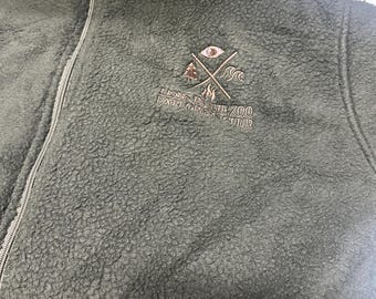 May include: An olive green fleece zip-up jacket featuring a high collar and a central zipper. Embroidered on the chest is a logo with crossed lines, an eye, a tree, a flame, and waves, with the text LIONS IN THE ZOO EXPLORERS CLUB.