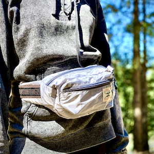 Op de afbeelding: Een lichtgrijze heuptas met een zwarte rits en een bruine band. De band heeft de tekst "SUN TREK" erop gedrukt. De heuptas heeft een klein patch met de tekst "CLONE" en een afbeelding van een vlinder.
