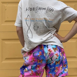 Pu&ograve; includere: Una persona indossa una maglietta beige chiaro con il testo "Lion in the Zoo" e un design grafico. La persona indossa anche pantaloncini colorati con una stampa di farfalla, fungo e pistola.