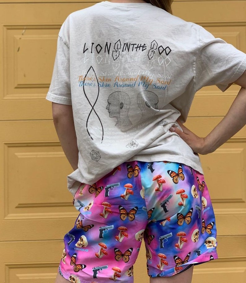 Pu&ograve; includere: Una persona indossa una maglietta beige chiaro con il testo "Lion in the Zoo" e un design grafico. La persona indossa anche pantaloncini colorati con una stampa di farfalla, fungo e pistola.