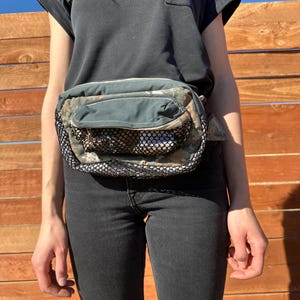 May include: A camouflage fanny pack with a black mesh pocket and a zipper closure. The fanny pack is worn around the waist of a person wearing a black t-shirt and black jeans.