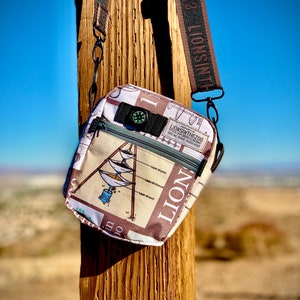 May include: A small, white and brown crossbody bag with a compass and the text "LIONSINTHEZOO" printed on it. The bag has a brown strap with the text "LIONSINTHEZOO" printed on it.