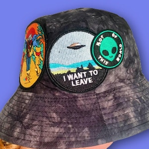 Könnte beinhalten: Ein schwarzer Bucket Hat mit Batik-Muster und verschiedenen gestickten Patches. Die Patches beinhalten ein UFO, einen grünen Außerirdischen und einen Patch mit der Aufschrift "Not of this world". Ein weiterer Patch lautet "I want to leave".