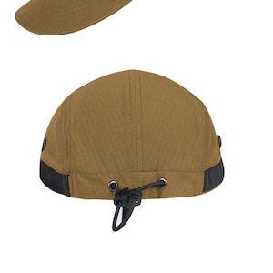 Puede incluir: Una gorra de béisbol de lona marrón con visera negra y correa ajustable. La gorra tiene dos ojales negros en la parte superior y una correa negra con hebilla en la parte posterior.