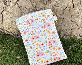 Op de afbeelding: Een etui met bloemenprint en de witte tekst "BOBBIN TAILS". De etui heeft een patroon van roze, gele en blauwe bloemen op een witte achtergrond. De etui ligt op groen gras en staat naast een boomstam.