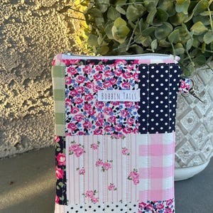 May include: A patchwork zippered pouch with a variety of floral, polka dot, and gingham patterns in pink, blue, and white. The pouch has a silver zipper and a small metal loop. The label on the pouch reads "BOBBIN TAILS".