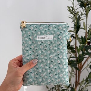 Pode incluir: Uma bolsa acolchoada verde menta com um padr&atilde;o floral branco e um z&iacute;per dourado. A bolsa tem um puxador de z&iacute;per em forma de estrela dourada e o texto "BOBBIN TAILS" na frente.