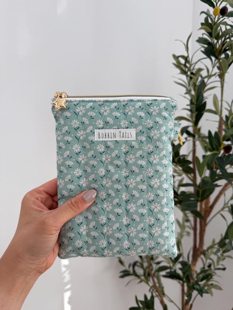 Pode incluir: Uma bolsa acolchoada verde menta com um padr&atilde;o floral branco e um z&iacute;per dourado. A bolsa tem um puxador de z&iacute;per em forma de estrela dourada e o texto "BOBBIN TAILS" na frente.