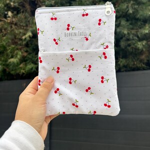 Pode incluir: Bolsa de tecido branco com padr&atilde;o de cerejas. A bolsa tem um pequeno bolso na frente e um fecho de correr. O texto "BOBBIN TAILS" est&aacute; impresso na frente. As cerejas s&atilde;o vermelhas com caules e folhas verdes.
