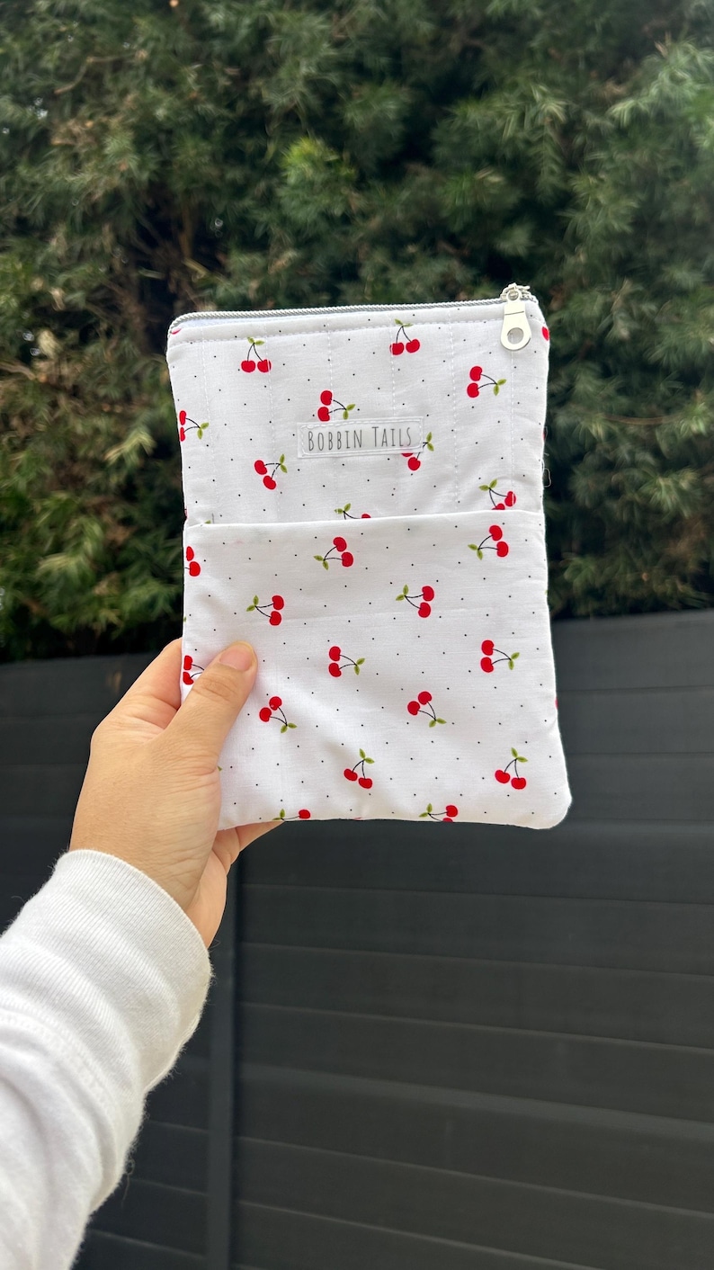Pode incluir: Bolsa de tecido branco com padr&atilde;o de cerejas. A bolsa tem um pequeno bolso na frente e um fecho de correr. O texto "BOBBIN TAILS" est&aacute; impresso na frente. As cerejas s&atilde;o vermelhas com caules e folhas verdes.