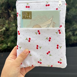 Pode incluir: Bolsa de tecido branco com estampa de cerejas, contendo um cart&atilde;o de felicita&ccedil;&otilde;es. A bolsa tem fecho de correr e aba. O cart&atilde;o de felicita&ccedil;&otilde;es tem um design decorativo e as palavras "Warm Greeting Card".