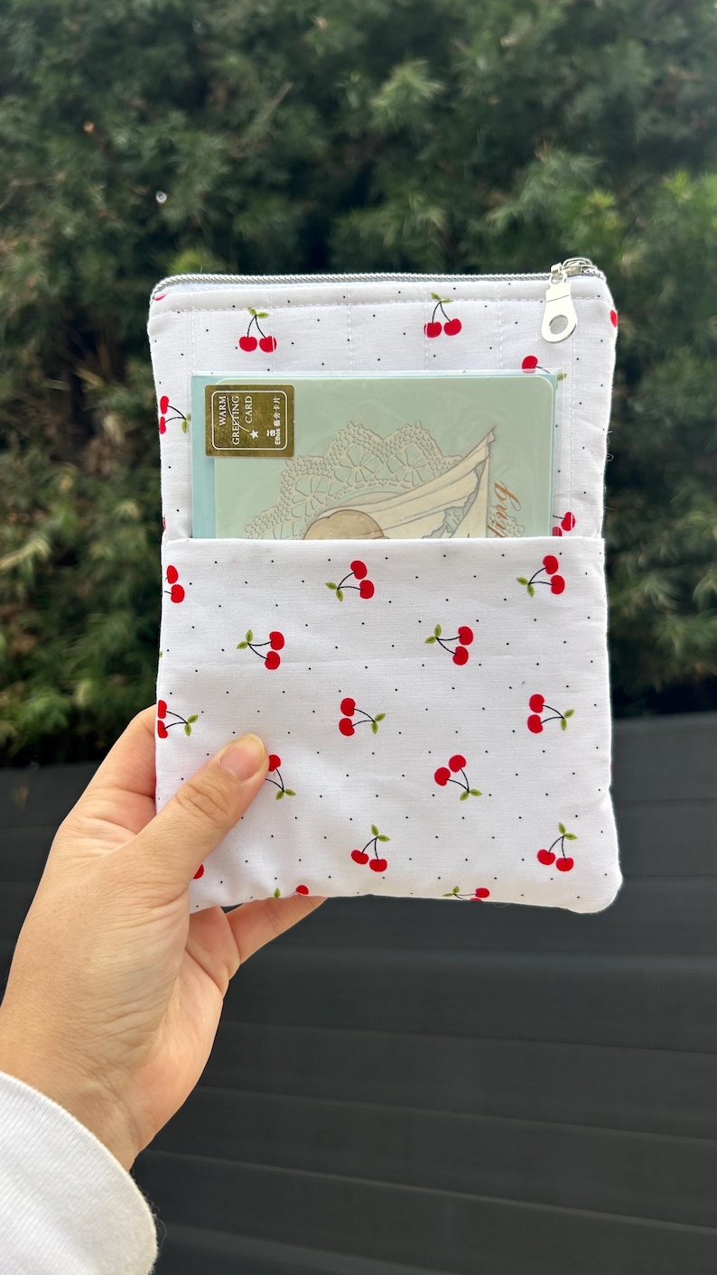 Pode incluir: Bolsa de tecido branco com estampa de cerejas, contendo um cart&atilde;o de felicita&ccedil;&otilde;es. A bolsa tem fecho de correr e aba. O cart&atilde;o de felicita&ccedil;&otilde;es tem um design decorativo e as palavras "Warm Greeting Card".