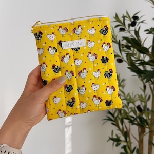Pode incluir: Bolsa acolchoada amarela com um padrão repetido de galinhas brancas e pretas com detalhes vermelhos. A bolsa tem um zíper dourado e o texto "BOBBIN TAILS" em uma etiqueta branca.