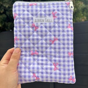 Pode incluir: Uma bolsa acolchoada com um padrão xadrez lavanda e branco, adornada com laços rosa. A bolsa possui um zíper prateado e o texto "BOBBIN TAILS". O item é segurado por uma pessoa.