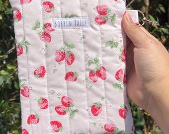 May include: A quilted, rectangular pouch with a zipper closure. The light pink fabric is patterned with red strawberries and green leaves. The text "BOBBIN TAILS" is printed on a white label. A small gold-colored ring is attached to the side.