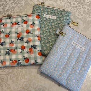 May include: Three fabric zipper pouches with gold-tone hardware. One pouch has a peach and green gingham pattern, another has a small white floral pattern on a green background, and the third has a small white floral pattern on a light blue background. Each pouch has a label that reads "BOBBIN TAILS".