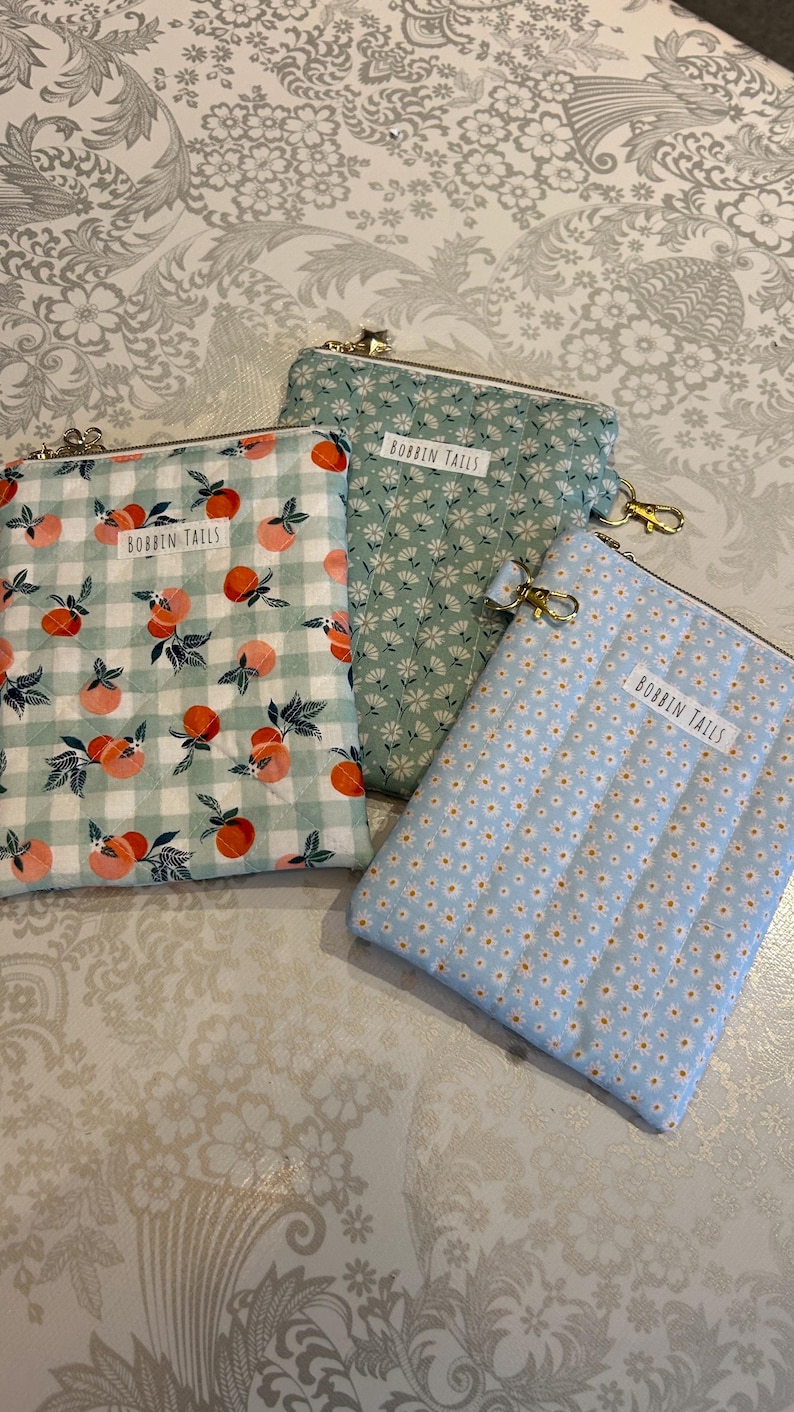 May include: Three fabric zipper pouches with gold-tone hardware. One pouch has a peach and green gingham pattern, another has a small white floral pattern on a green background, and the third has a small white floral pattern on a light blue background. Each pouch has a label that reads "BOBBIN TAILS".