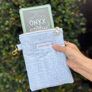 May include: A light blue e-reader sleeve with a daisy pattern, gold zip, and a flower charm. The text "BOBBIN TAILS" is visible on the front. An e-reader with the text "ONYX STORM" is partially visible inside the sleeve.