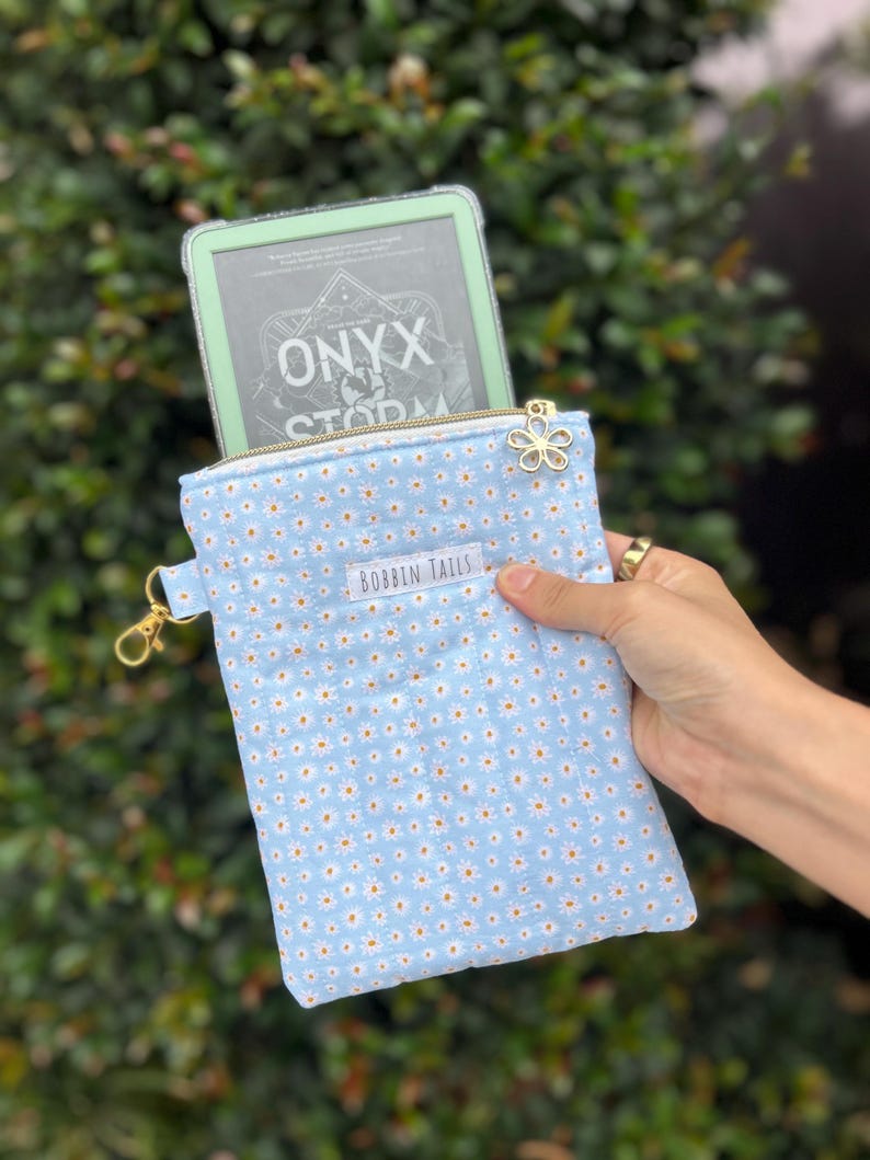 May include: A light blue e-reader sleeve with a daisy pattern, gold zip, and a flower charm. The text "BOBBIN TAILS" is visible on the front. An e-reader with the text "ONYX STORM" is partially visible inside the sleeve.