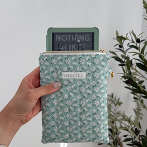 May include: A mint green e-reader case with a white floral pattern and a gold zip. The case has a label that reads "BOBBIN TAILS". The e-reader displays the text "NOTHING LIKE".