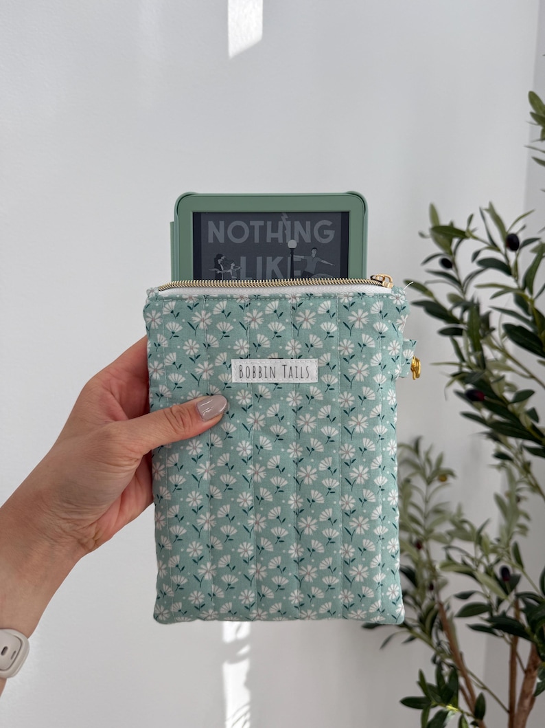 Pode incluir: Um e-reader verde-menta &eacute; parcialmente vis&iacute;vel dentro de uma capa com estampa floral. A capa tem um padr&atilde;o de margaridas brancas em um fundo verde-menta, um z&iacute;per dourado e o texto "BOBBIN TAILS". O e-reader exibe o texto "NOTHING LIKE".