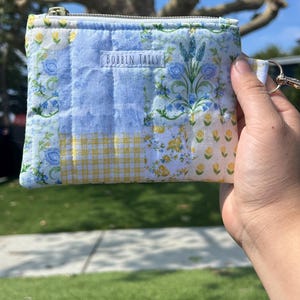 Könnte beinhalten: Ein gesteppter Beutel mit Blumen- und Vichy-Muster in Blau-, Gelb- und Weißtönen. Der Beutel hat einen Reißverschluss und eine kleine Schlaufe mit einem Metallverschluss. Der Text "BOBBIN TALLS" ist auf der Vorderseite aufgedruckt.