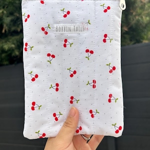 Puede incluir: Bolsa acolchada blanca con un patr&oacute;n repetido de cerezas rojas y tallos verdes. La bolsa tiene una cremallera plateada y el texto "BOBBIN TAILS" est&aacute; impreso en la parte delantera.