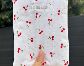 May include: White quilted pouch with a repeating pattern of red cherries and green stems. The pouch has a silver zipper and the text "BOBBIN TAILS" is printed on the front.