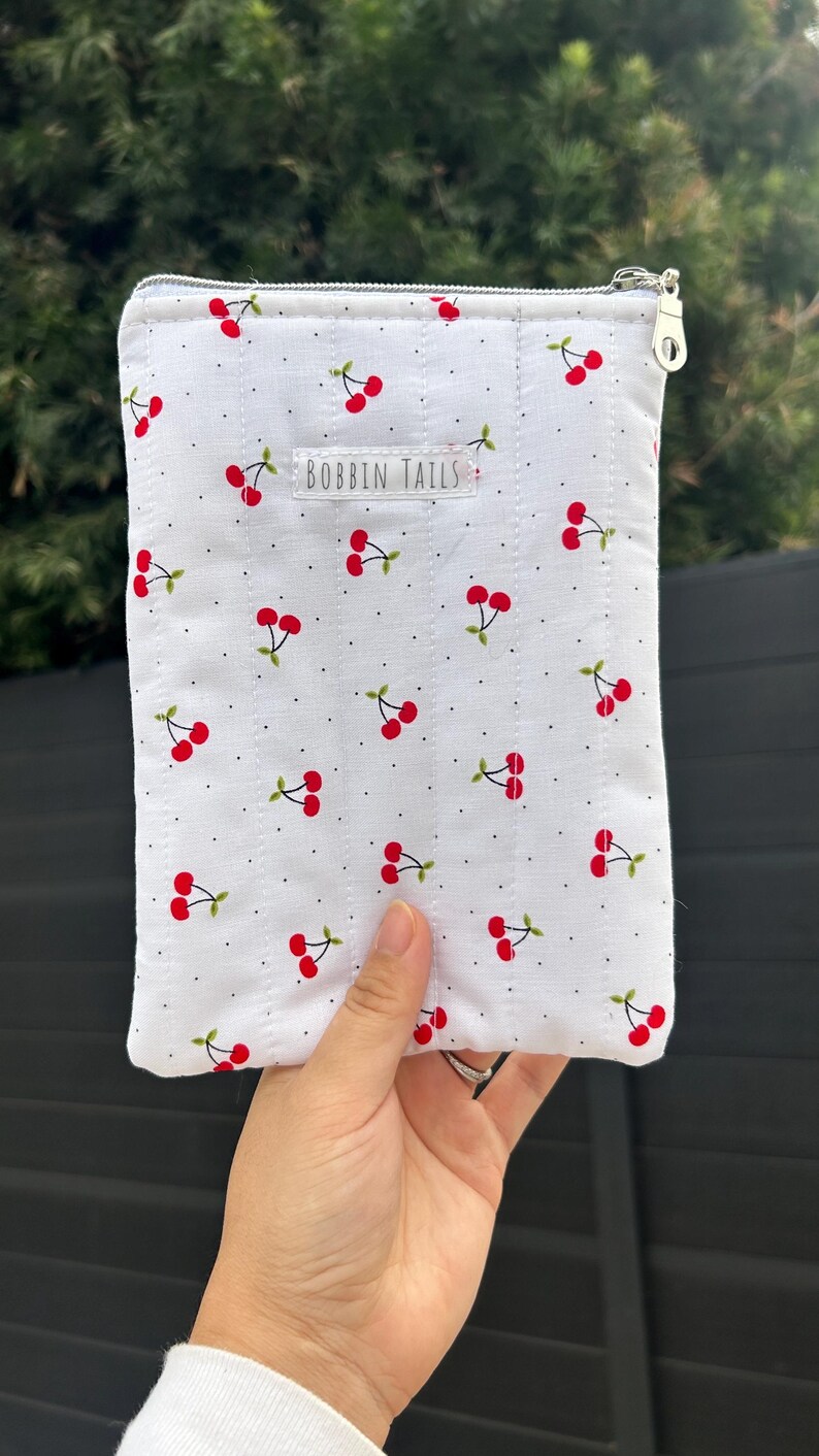 Puede incluir: Bolsa acolchada blanca con un patr&oacute;n repetido de cerezas rojas y tallos verdes. La bolsa tiene una cremallera plateada y el texto "BOBBIN TAILS" est&aacute; impreso en la parte delantera.