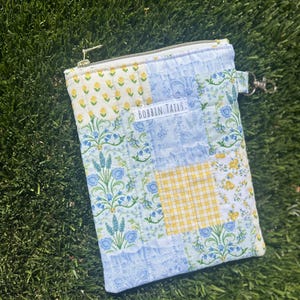 May include: A quilted pouch with a patchwork design featuring floral and gingham patterns in shades of blue, yellow, and white. The pouch has a zip closure and a small tag that reads "BOBBIN TAILS".