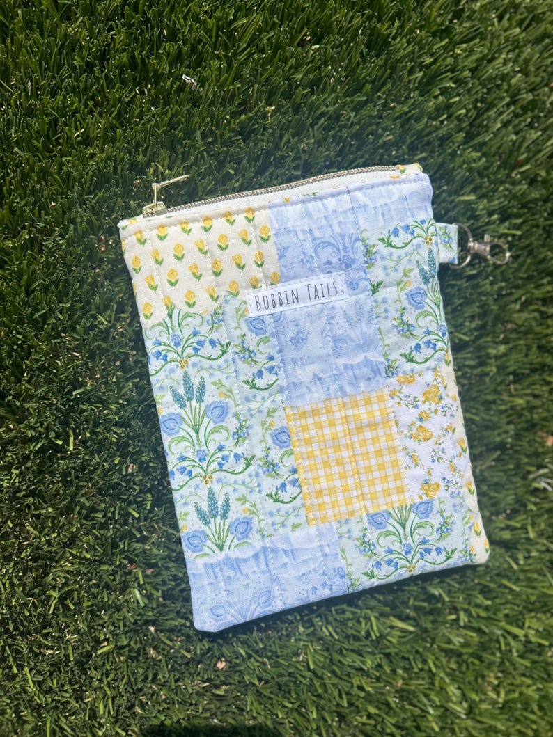 May include: A quilted pouch with a patchwork design featuring floral and gingham patterns in shades of blue, yellow, and white. The pouch has a zip closure and a small tag that reads "BOBBIN TAILS".
