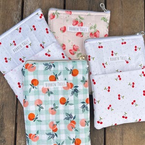 Pode incluir: Uma cole&ccedil;&atilde;o de bolsas com z&iacute;per com v&aacute;rios padr&otilde;es de frutas, incluindo cerejas e p&ecirc;ssegos, em uma superf&iacute;cie de madeira. As bolsas s&atilde;o em tons de branco, rosa e verde, com o texto "BOBBIN TAILS" em cada uma.