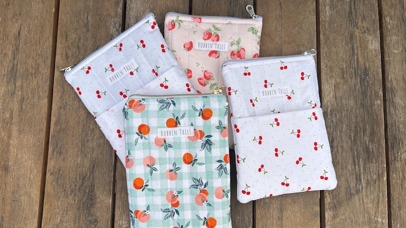 Pode incluir: Uma cole&ccedil;&atilde;o de bolsas com z&iacute;per com v&aacute;rios padr&otilde;es de frutas, incluindo cerejas e p&ecirc;ssegos, em uma superf&iacute;cie de madeira. As bolsas s&atilde;o em tons de branco, rosa e verde, com o texto "BOBBIN TAILS" em cada uma.
