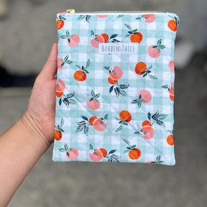 Pode incluir: Uma bolsa acolchoada com um padrão xadrez vichy azul claro e branco, adornada com ilustrações de pêssegos. A bolsa tem um zíper dourado e o texto "BOBBIN TAILS" em uma etiqueta branca. A bolsa é segurada em uma mão.