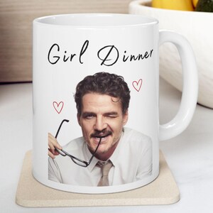 May include: White ceramic mug with the text "Girl Dinner" in black script, featuring a photo of a man holding glasses. The mug has three small red heart illustrations. The mug is on a beige coaster.